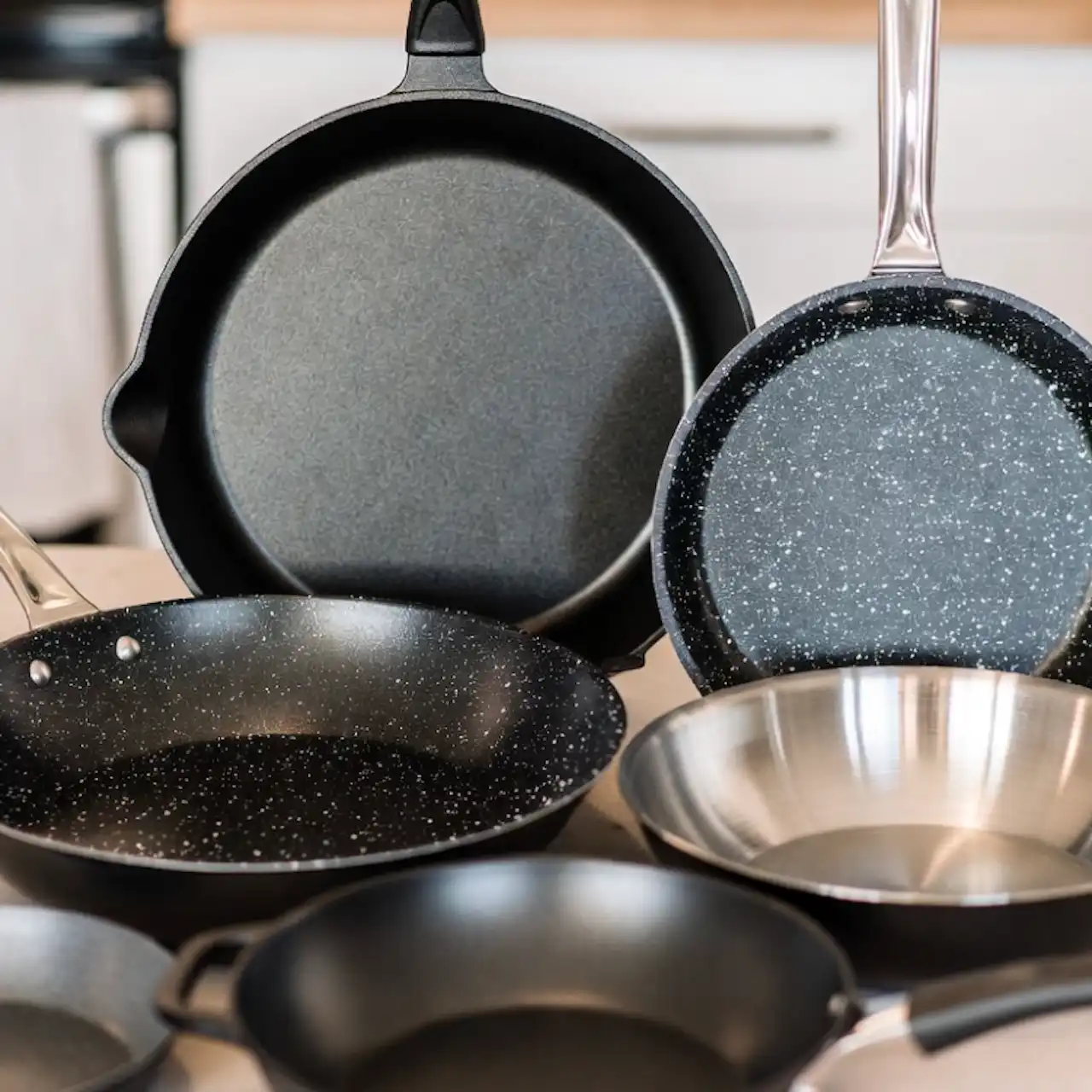 Diferentes tipos de sartenes (hierro, cerámica, acero) dispuestas sobre una mesa de cocina.
